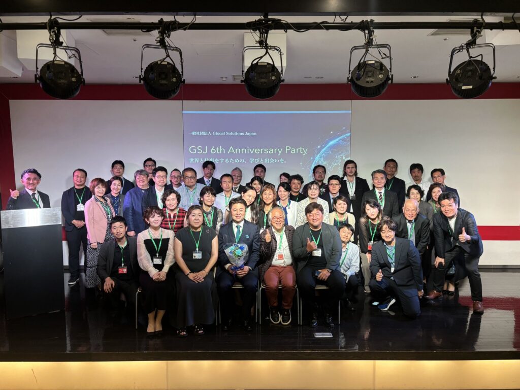 GSJ 6周年記念パーティーの集合写真。ステージのスクリーンに「GSJ 6th Anniversary Party」と映し出され、その前に参加者が並んでいる