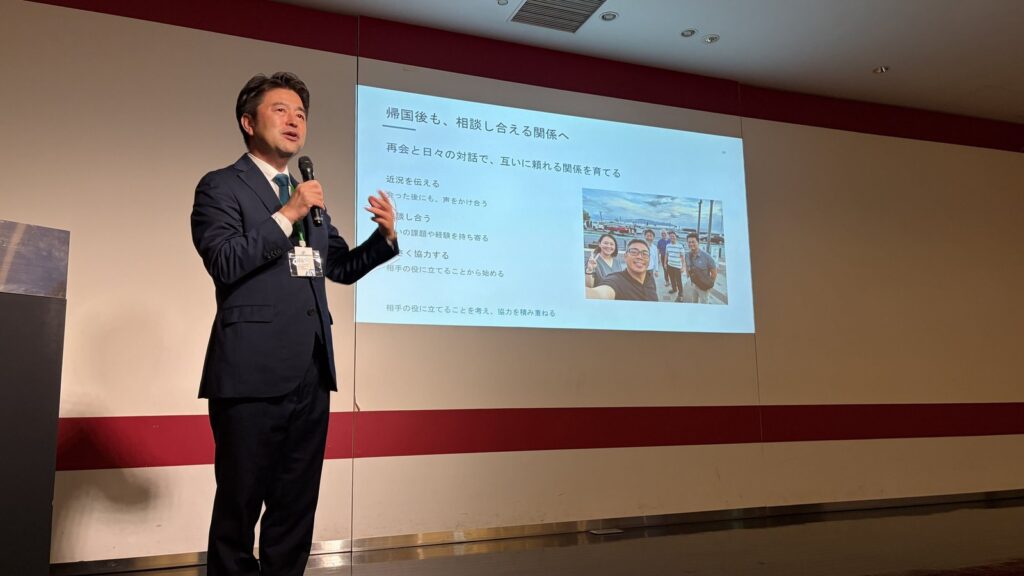 スクリーンの前でマイクを持って話す深野裕之代表理事